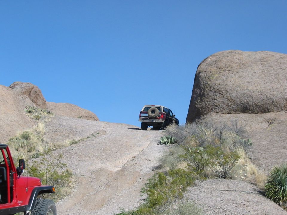 Red Jeep and a white and brown Ford Bronco driving up a dirt hill in a rocky, desert landscape
