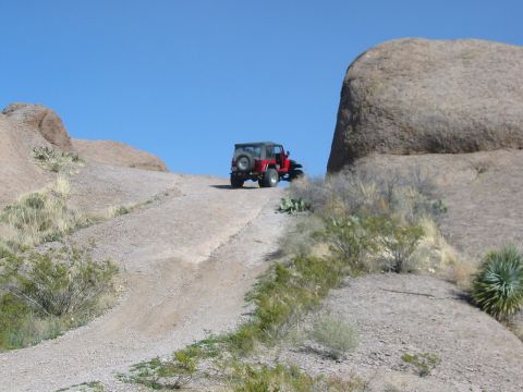 1996 Ford Bronco - A red Jeep Wrangler navigates a rocky, off-road trail under a clear blue sky.