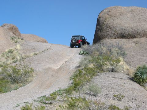 1996 Ford Bronco - A red Jeep Wrangler navigates a steep, gravelly path in a desert landscape with large rock formations.