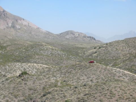 1996 Ford Bronco - A lone red SUV traverses a dusty track through a desolate, mountainous terrain under a clear blue sky.