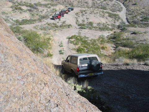 1996 Ford Bronco - Multiple SUVs, including a tan Ford Bronco, navigate a winding dirt path in a desert landscape.
