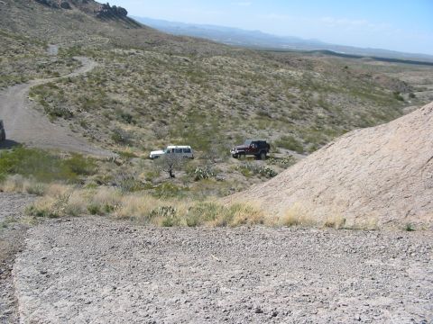1996 Ford Bronco - Two off-road vehicles navigate a dusty trail through a barren, mountainous terrain under a clear blue sky.