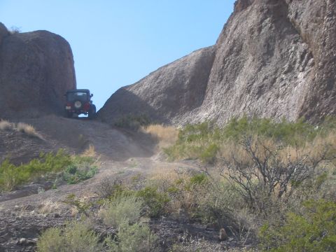 1996 Ford Bronco - A red Jeep Wrangler navigates a dusty trail in a rocky, arid landscape.