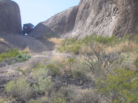 1996 Ford Bronco - A red off-road vehicle navigates a steep, rocky path between large rock formations under a clear blue sky.