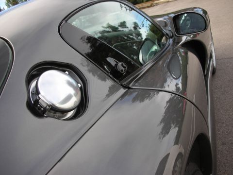 1998 Ford Crown Victoria - Close-up of a gray sports car's polished round fuel-filler cap beside the side window and mirror, trees reflected in the paint.