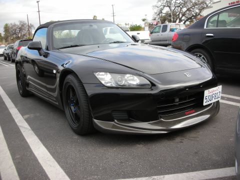 1998 Ford Crown Victoria - Front three-quarter of a black Honda S2000 roadster with black wheels and a front splitter, parked in a tree-lined lot.