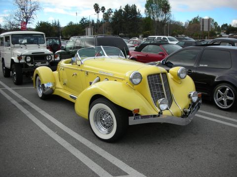 1998 Ford Crown Victoria - yellow Auburn-style boattail speedster roadster with whitewall wire wheels and chrome trim parked beside a white FJ40 and SUVs.