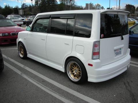 1998 Ford Crown Victoria - Rear three-quarter of a lowered white Scion xB box-wagon on gold split-spoke wheels with window visors, parked at a car meet.