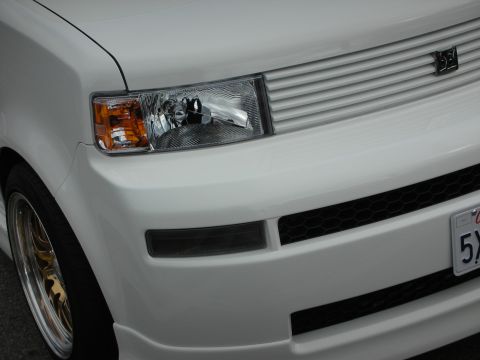 1998 Ford Crown Victoria - Close-up of a white Scion xB's front corner showing the clear headlight, lower grille and a small badge on the fender.