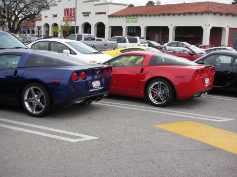 1998 Ford Crown Victoria - blue and a red Chevrolet Corvette coupe parked rear-to-rear in a shopping-center lot, both on polished wheels with round taillights.