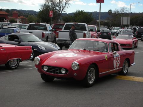 1998 Ford Crown Victoria - red vintage Ferrari coupe carrying race number 155 rolls through a parking lot past other classic cars.