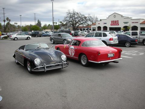 1998 Ford Crown Victoria - black Porsche 356 Speedster parked nose-to-nose with a red Ferrari coupe bearing number 155 in a Vons shopping center lot.