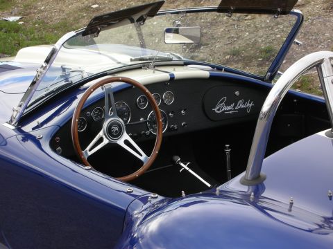 1998 Ford Crown Victoria - cockpit of a blue Shelby Cobra roadster, showing a wood-rimmed steering wheel, gauges, and a Carroll Shelby dash signature.