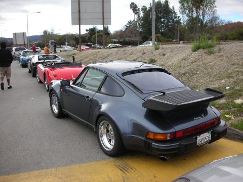 1998 Ford Crown Victoria - gray Porsche 911 Turbo with its signature whale-tail spoiler, parked in a roadside line of classic cars.