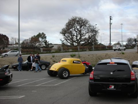 1998 Ford Crown Victoria - bright yellow fenderless hot rod coupe is parked among other cars and onlookers in a lot under cloudy skies.