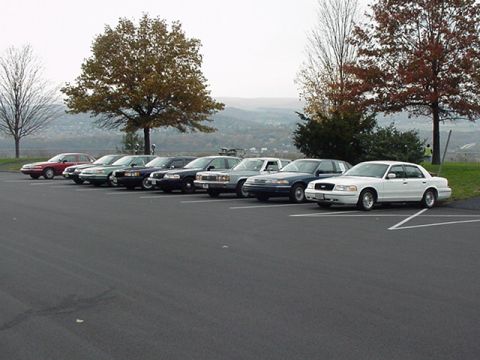 1989 Ford LTD Crown Victoria - A row of full-size sedans, likely Ford Crown Victorias, parked diagonally in a paved lot with fall trees and hills in the background. The c…