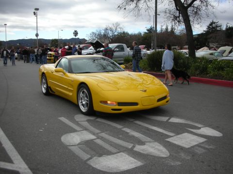 1998 Ford Crown Victoria - yellow Chevrolet Corvette coupe sits over a faded crosswalk as crowds gather among cars in the background.
