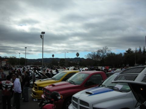 1998 Ford Crown Victoria - Rows of cars and trucks fill a car-show parking lot under heavy gray clouds with hills behind.