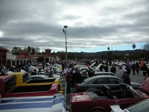 1998 Ford Crown Victoria - wide view of a packed car meet with dozens of cars, raised hoods, and crowds beneath overcast skies.