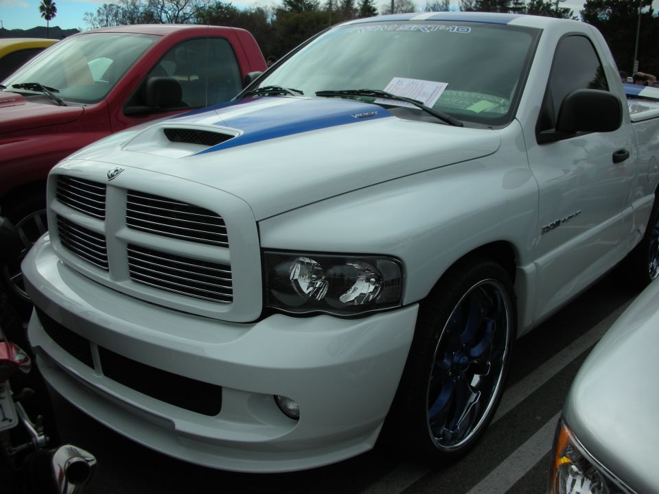 View over a car-show lot through bare tree branches with a silver Cadillac Escalade