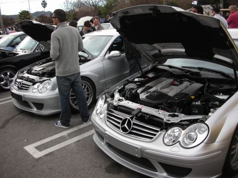 1998 Ford Crown Victoria - Two silver Mercedes-Benz CLK coupes sit with hoods raised at a car meet while a man stands between them.
