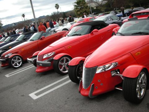 1998 Ford Crown Victoria - row of red Plymouth Prowler roadsters with open-wheel front ends lines up among cars at an outdoor show.
