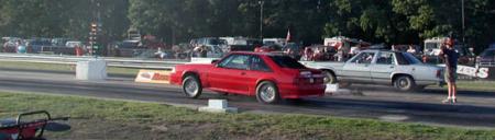 1989 Ford LTD Crown Victoria - A red Ford Mustang (Fox-body) coupe launches down a drag strip, shown in side/3q-rear view with its front end slightly raised, while a whit…