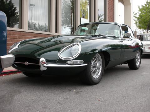 1998 Ford Crown Victoria - dark green Jaguar E-Type coupe parked along a brick building, showing its long hood, oval grille and chrome wire wheels.