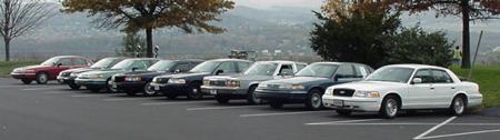 1989 Ford LTD Crown Victoria - A row of full-size sedans, likely Ford Crown Victorias, parked diagonally in a lot overlooking a hazy valley. Colors range from maroon and…