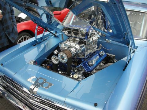 1998 Ford Crown Victoria - chrome-topped supercharged V8 sits in the engine bay of a light-blue classic car with its hood raised at a parking-lot show.