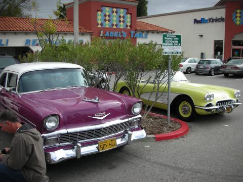 1998 Ford Crown Victoria - purple mid-1950s Chevrolet and a yellow Corvette roadster parked outside Label's Table and FedEx Kinko's storefronts.