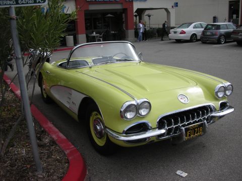 1998 Ford Crown Victoria - yellow late-1950s Chevrolet Corvette convertible with chrome grille and dual headlights parked at a strip-mall curb.