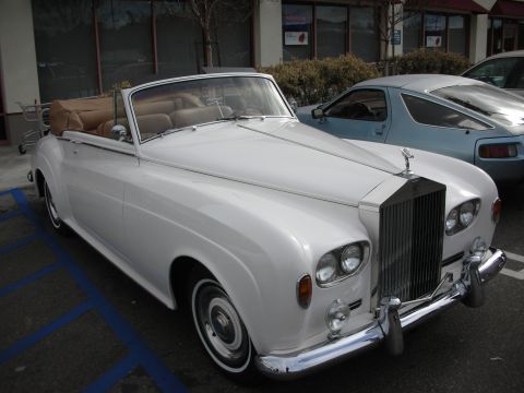 1998 Ford Crown Victoria - white Rolls-Royce drophead convertible with tan interior and top down parked beside a blue coupe in a lot.