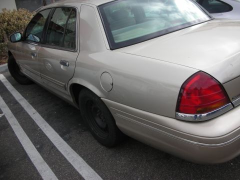 1998 Ford Crown Victoria - tan Ford Crown Victoria sedan with black steel wheels shown from the rear three-quarter in marked parking spaces.