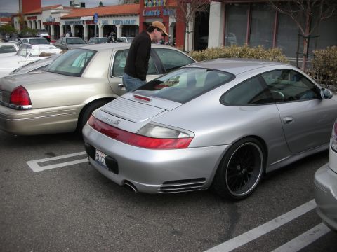 1998 Ford Crown Victoria - silver Porsche 911 Carrera coupe with black wheels parked behind a tan sedan as a man walks past at a strip mall.