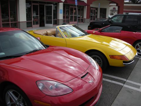1998 Ford Crown Victoria - red Chevrolet Corvette C6 parked nose-out next to a yellow Ferrari Daytona-style Spyder outside storefronts.