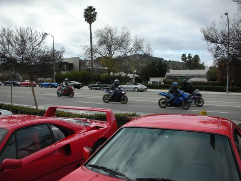 1998 Ford Crown Victoria - Riders on blue and dark motorcycles pass along a road behind parked red sports cars under an overcast sky.