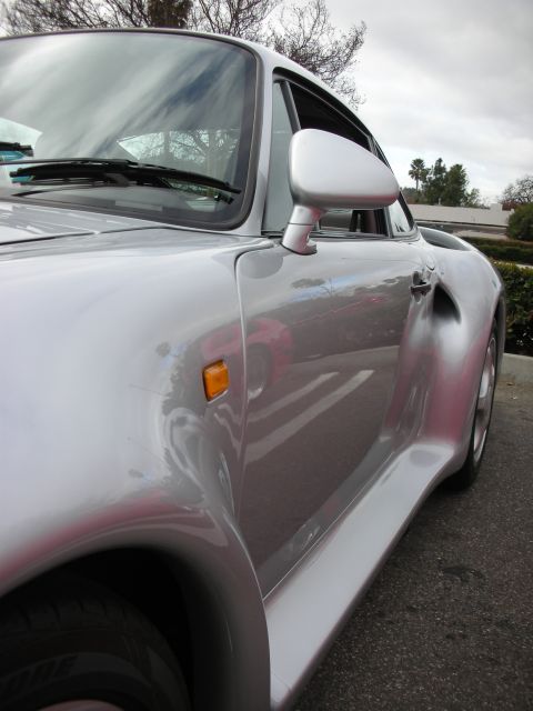 1998 Ford Crown Victoria - close-up of a silver Porsche's side mirror, door, orange marker light and rear fender flare under a cloudy sky.