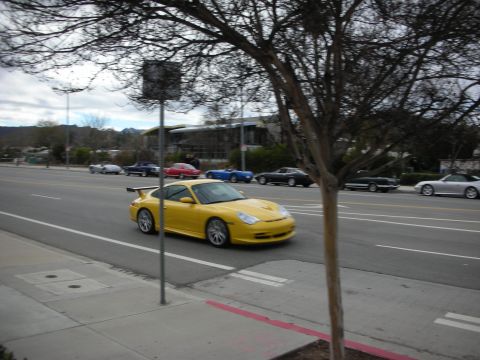 1998 Ford Crown Victoria - yellow Porsche 911 GT3 with a tall rear wing drives along a street past parked cars under a gray sky.