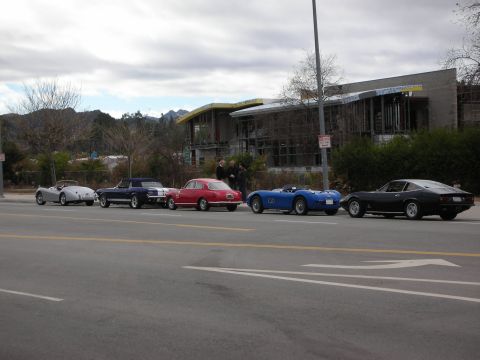 1998 Ford Crown Victoria - lineup of vintage roadsters and a dark coupe parked along a roadside in front of a modern building under gray skies.