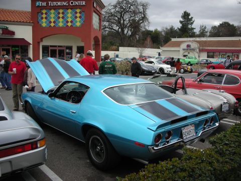 1998 Ford Crown Victoria - light-blue early-1970s Chevrolet Camaro with black hood stripes and open hood parked among cars at a strip-mall meet.
