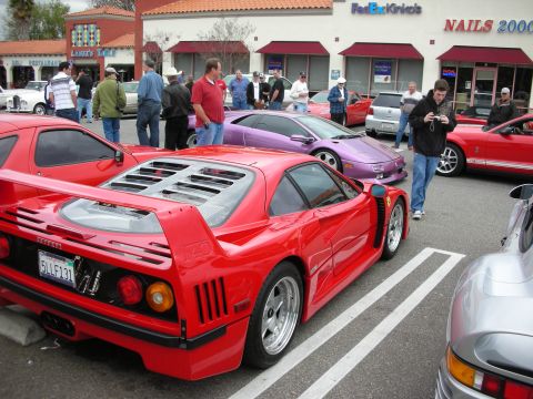 1998 Ford Crown Victoria - red Ferrari F40 sits in a parking-lot car meet, its slatted engine cover and rear wing facing a crowd of spectators and storefront signs.