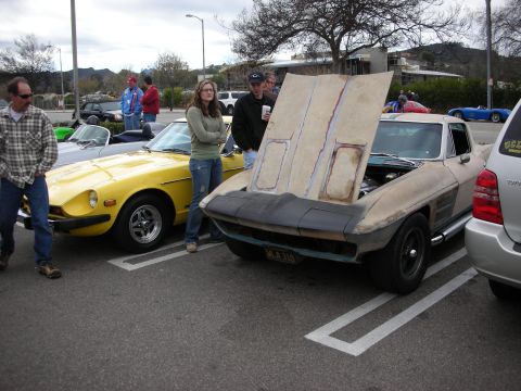 1998 Ford Crown Victoria - silver Chevrolet Corvette sits with its primer-gray front clamshell tilted up, parked beside a yellow Datsun Z and spectators.