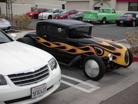 1998 Ford Crown Victoria - black hot-rod coupe with orange-and-yellow flame paint and exposed front suspension is parked next to a white Chrysler Crossfire.