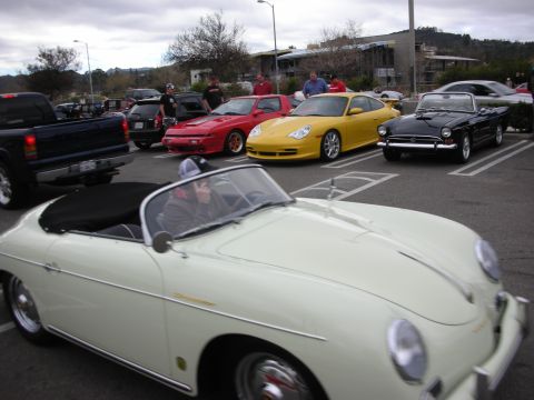 1998 Ford Crown Victoria - cream Porsche 356 Speedster drives through a parking lot past a row of parked sports cars under an overcast sky.