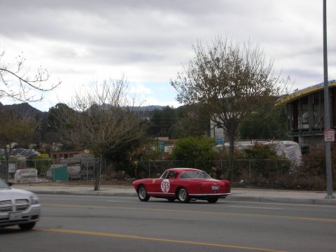1998 Ford Crown Victoria - red vintage Italian coupe wearing a circular race number drives along a roadside lined by a chain-link fence and bare trees.