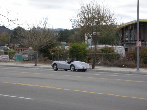 1998 Ford Crown Victoria - small silver vintage roadster with cut-down doors drives along a roadside with bare trees and distant hills in the background.