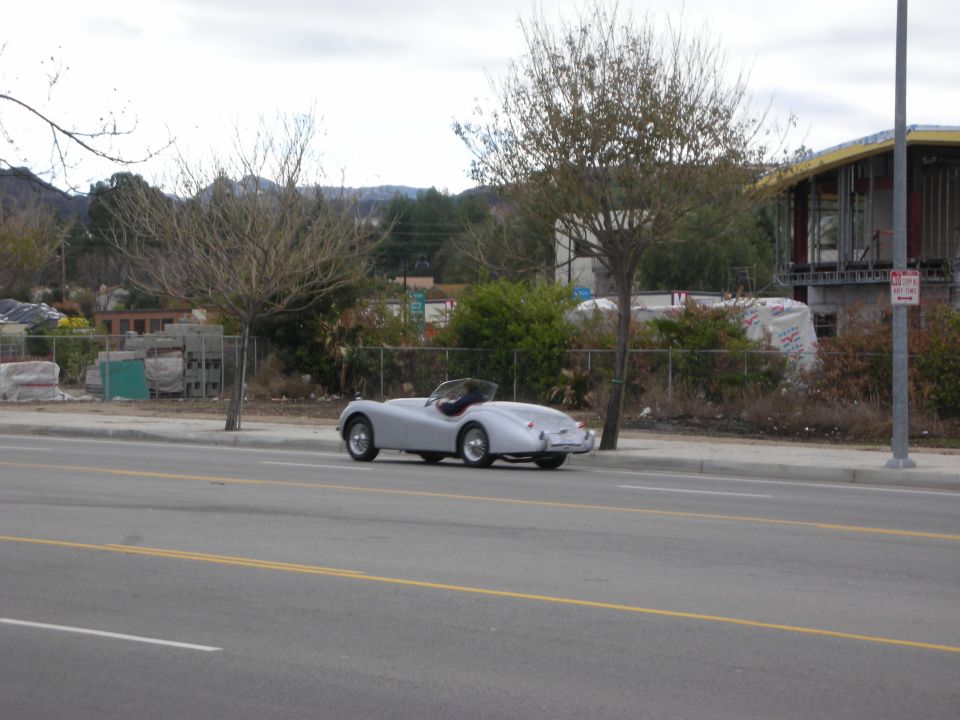 Silver vintage roadster driving on a road beside a chain-link fence and hills