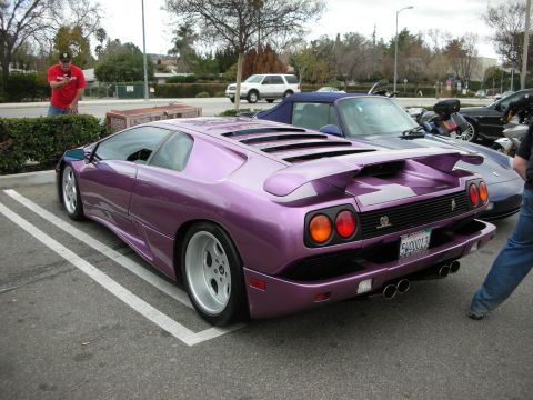 1998 Ford Crown Victoria - purple Lamborghini Diablo sits angled in a parking space with its rear wing raised as onlookers stand nearby.
