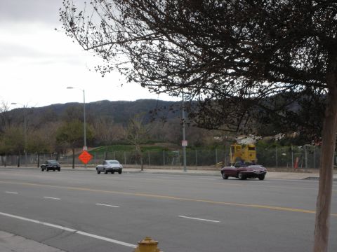 1998 Ford Crown Victoria - dark red convertible and other cars travel along a multi-lane road with construction equipment, a chain-link fence and hills behind.